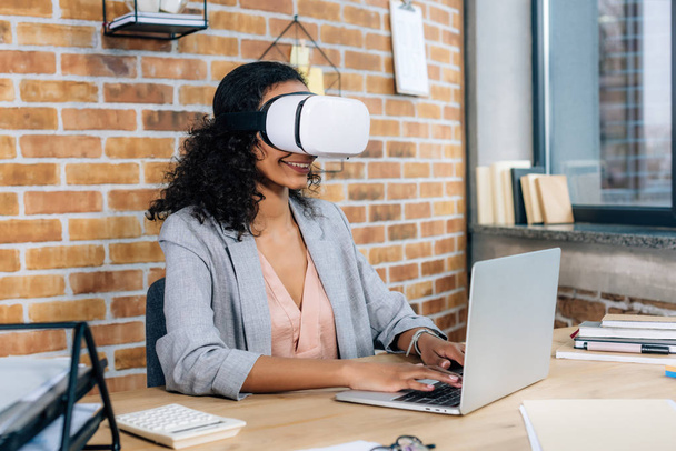 Afrikkalainen amerikkalainen rento liikenainen virtuaalitodellisuus kuulokkeet toimistopöytä kannettavan tietokoneen avulla
 - Valokuva, kuva