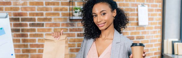 panoramic shot of african american Casual businesswoman holding takeaway paper bag and coffee to go in office - Foto, Bild