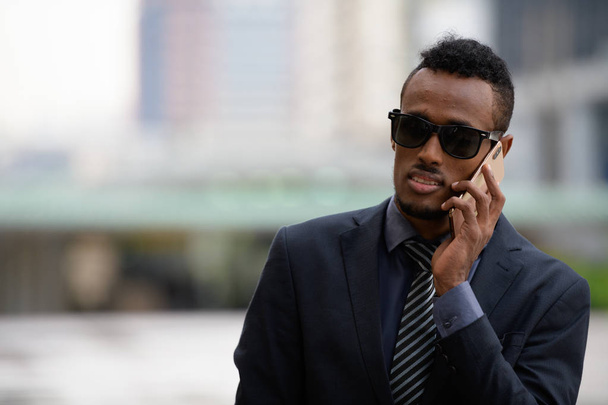 Nuori afrikkalainen liikemies, jolla on aurinkolasit, puhuu puhelimessa ulkona
 - Valokuva, kuva