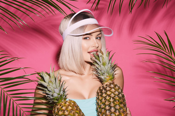 portrait of beautiful young woman in swimwear and cap posing with pineapples on pink background with palm leaves - Foto, Imagem