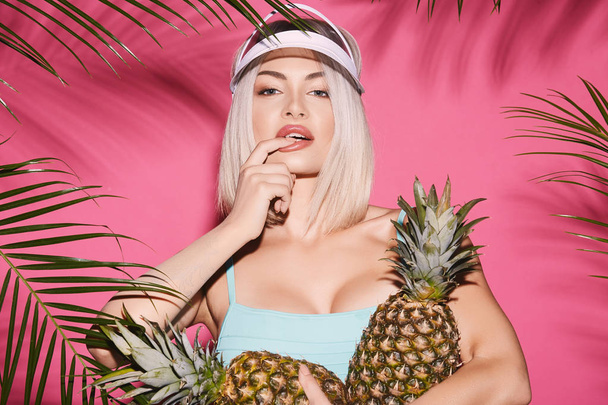 portrait of beautiful young woman in swimwear and cap posing with pineapples on pink background with palm leaves - Photo, Image
