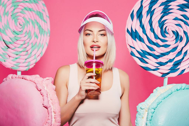 Attractive girl with blonde hair standing with fruits cocktail and huge sweets on pink background - Photo, Image