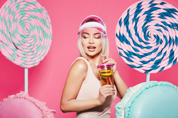 Приваблива дівчина з світлим волоссям, що стоїть з фруктовим коктейлем і величезними цукерками на рожевому фоні
 - Фото, зображення