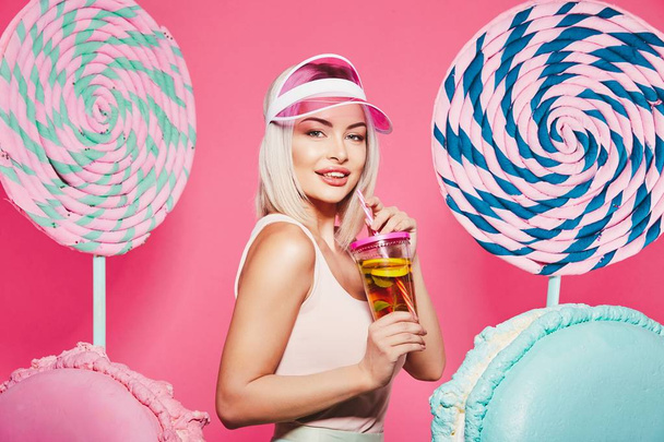 Atrakcyjna dziewczyna z blond włosy stojący z owocami koktajl i ogromne słodycze na różowym tle - Zdjęcie, obraz