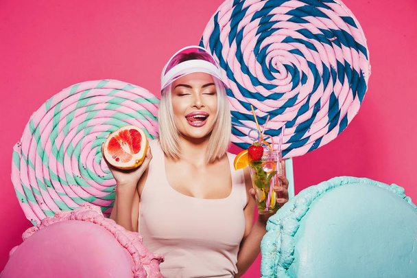 Aantrekkelijk meisje met blonde haar staande met vruchten cocktail en enorme snoepjes op roze achtergrond - Foto, afbeelding