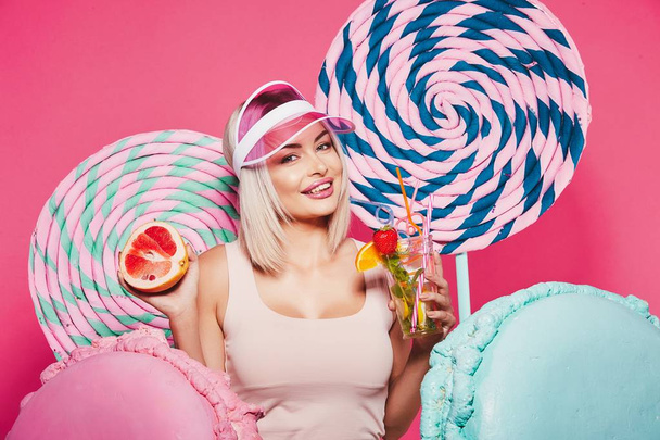 Attractive girl with blonde hair standing with fruits cocktail and huge sweets on pink background - Photo, Image