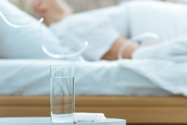 selective focus of sick senior woman lying on bed in clinic with glass of water on foreground - Photo, Image
