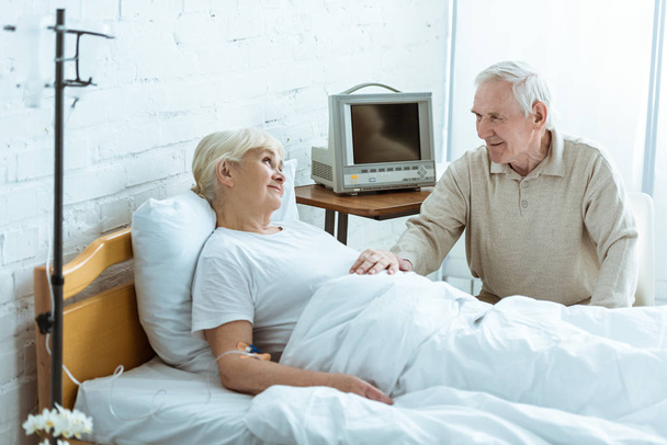 smiling sick senior woman looking at husband in clinic - Фото, изображение