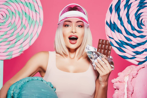 Attractive girl with blonde hair standing with chocolate and huge sweets on pink background - Photo, Image