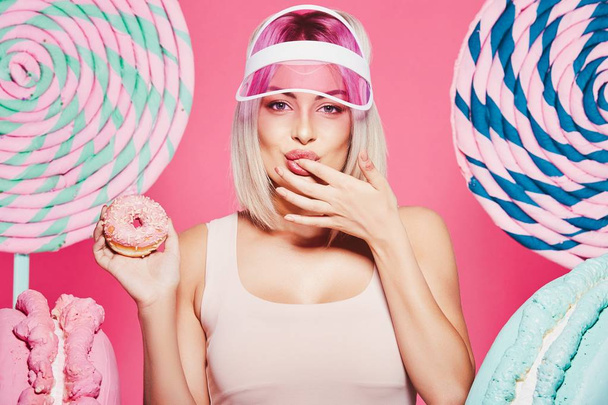 Atrakcyjna dziewczyna z blond włosami stojącą z pączką i ogromnymi słodyczami na różowym tle - Zdjęcie, obraz