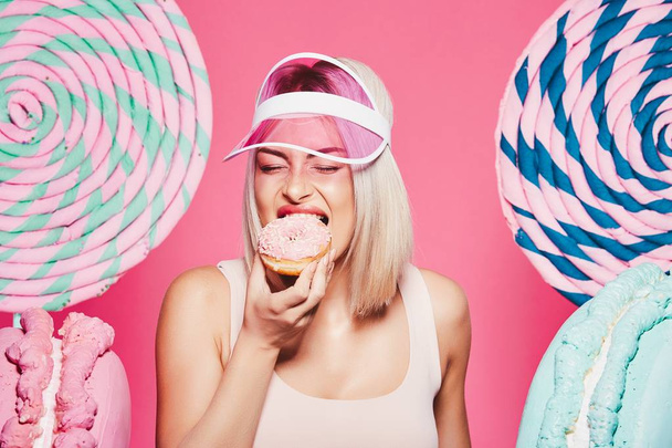 attraktives Mädchen mit blonden Haaren, stehend mit Donut und riesigen Süßigkeiten auf rosa Hintergrund - Foto, Bild