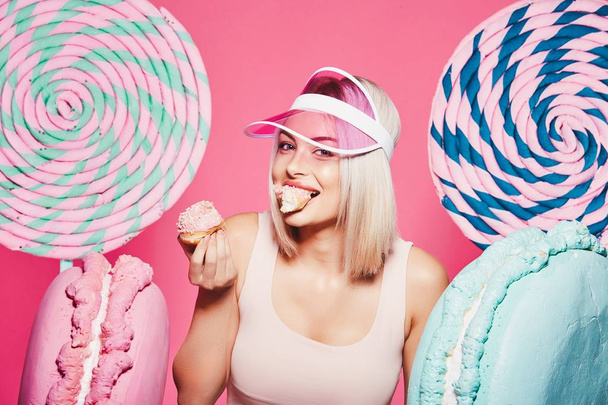 Attractive girl with blonde hair standing with doughnut and huge sweets on pink background - Foto, afbeelding