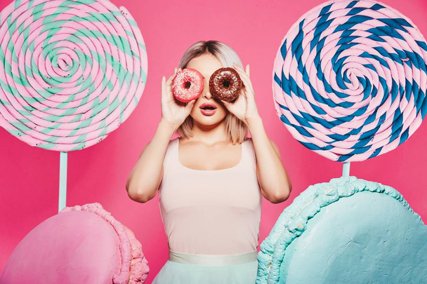 attraktives Mädchen mit blonden Haaren, stehend mit Donuts und riesigen Süßigkeiten auf rosa Hintergrund - Foto, Bild