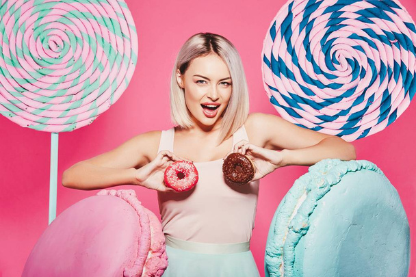 Attractive girl with blonde hair standing with doughnuts and huge sweets on pink background - Foto, Bild