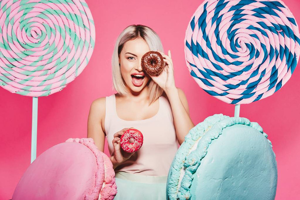 Aantrekkelijk meisje met blond haar staande met donuts en enorme snoepjes op roze achtergrond - Foto, afbeelding