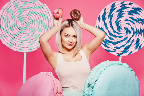 Приваблива дівчина з білявим волоссям, що стоїть з пончиками і величезними цукерками на рожевому фоні
 - Фото, зображення