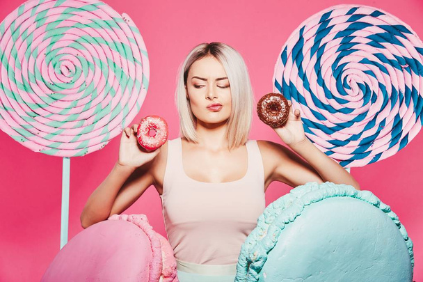 Привлекательная девушка с светлыми волосами, стоящая с пончиками и огромными сладостями на розовом фоне
 - Фото, изображение