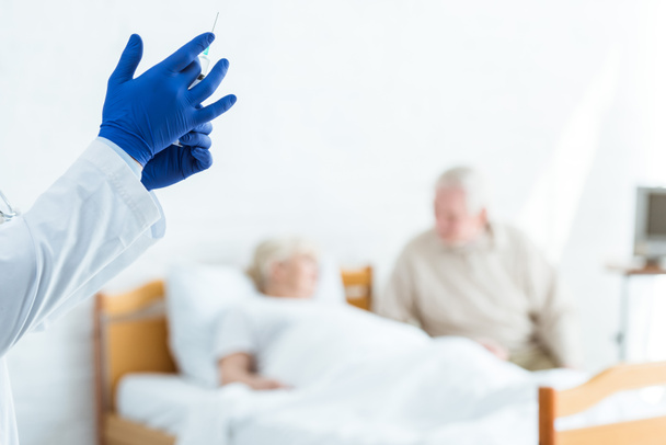 gedeeltelijke weergave van arts in latex handschoenen met spuit, zieke patiënt en senior man in de kliniek - Foto, afbeelding