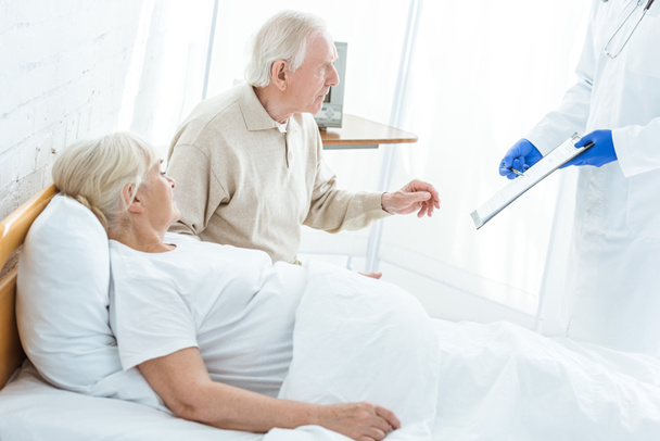 częściowy widok lekarza trzymania schowka, chorego pacjenta i starszego mężczyzny w klinice - Zdjęcie, obraz