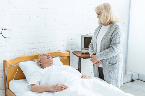 старша жінка стоїть біля хворого чоловіка в лікарні
 - Фото, зображення