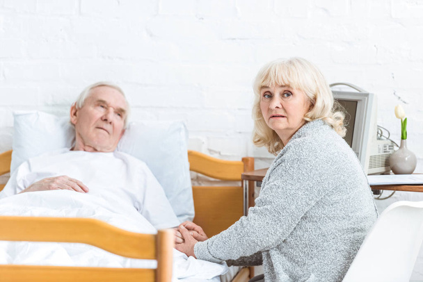 беспокойная пожилая женщина сидит рядом с больным мужем и держит его за руку в больнице
 - Фото, изображение