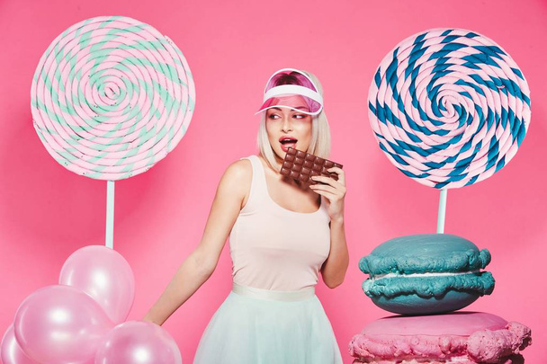 Menina atraente com cabelo loiro de pé com enormes doces no fundo rosa enquanto come chocolate
 - Foto, Imagem