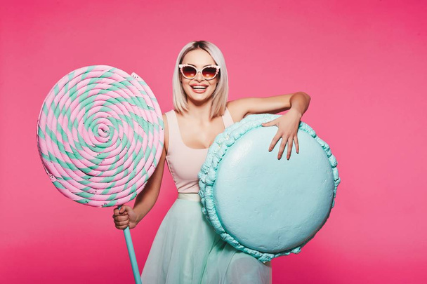 Attractive girl with blonde hair posing with huge lollipop and macaron on pink background - Foto, Imagen