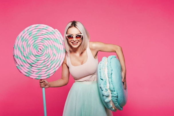 Aantrekkelijk meisje met blonde haren poseren met enorme Lollipop en Macaron op roze achtergrond - Foto, afbeelding