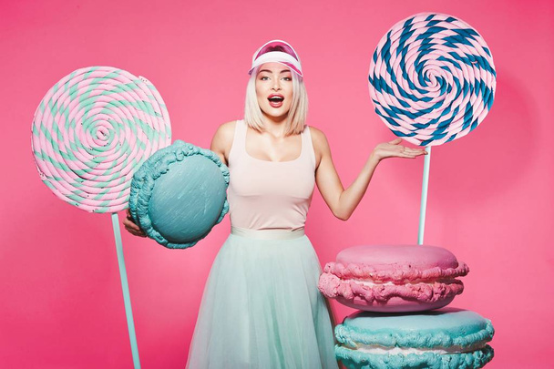 ピンクの背景に巨大なお菓子で立っているブロンドの髪を持つ魅力的な女の子 - 写真・画像