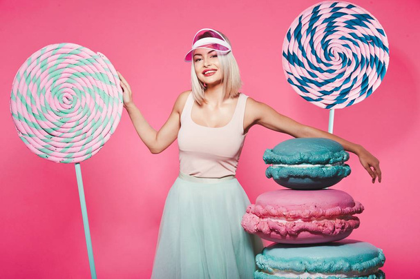 Attractive girl with blonde hair standing with huge sweets on pink background - Fotó, kép