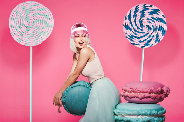 Muchacha atractiva con el pelo rubio de pie con enormes dulces sobre fondo rosa
 - Foto, imagen