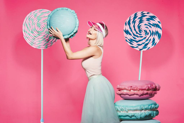 Muchacha atractiva con el pelo rubio de pie con enormes dulces sobre fondo rosa
 - Foto, Imagen
