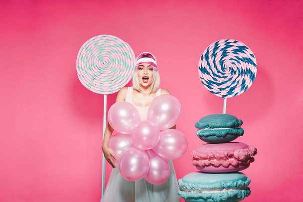 Aantrekkelijk meisje met blond haar staande met enorme snoepjes op roze achtergrond - Foto, afbeelding