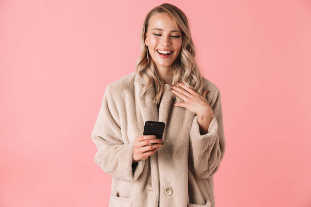 Laughing blonde woman wearing in fur coat holding smartphone - Photo, Image