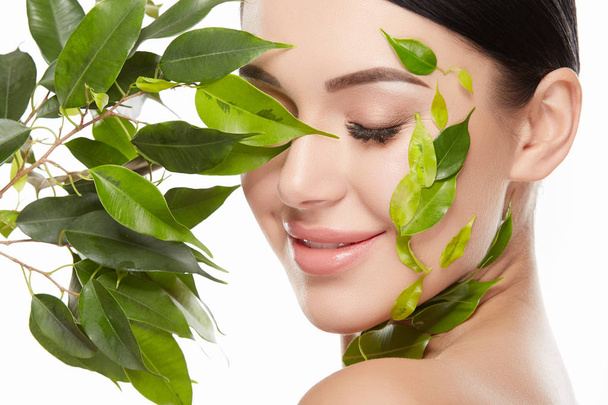 Porträt einer jungen Frau mit nacktem Make-up und grünen Blättern im Gesicht auf weißem Hintergrund - Foto, Bild