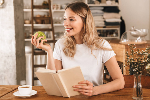 Portrait of smart blond woman reading book and eating green appl - 写真・画像
