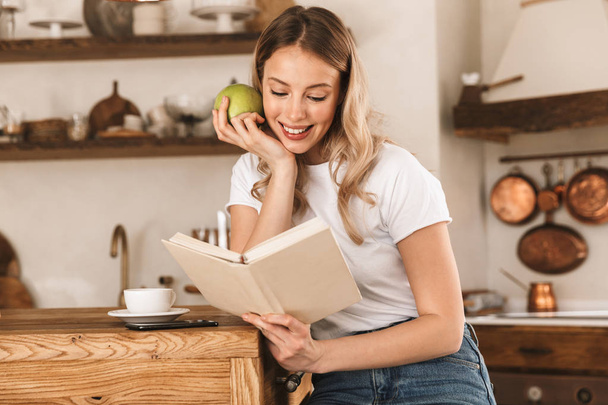 Portrait of relaxed blond woman reading book and eating green ap - Foto, Imagem