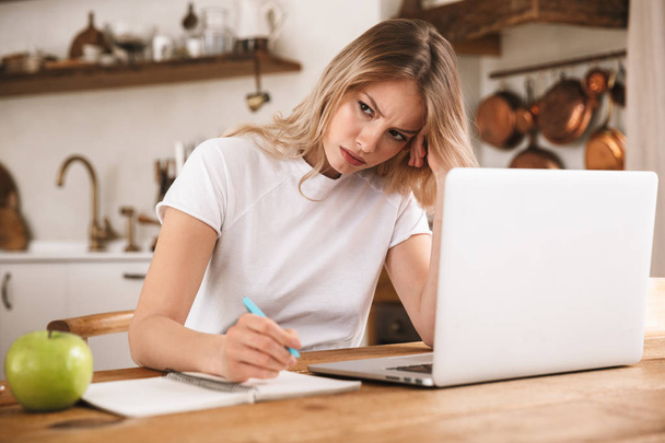 Image of sad blond woman studying on laptop and writing down not - Φωτογραφία, εικόνα