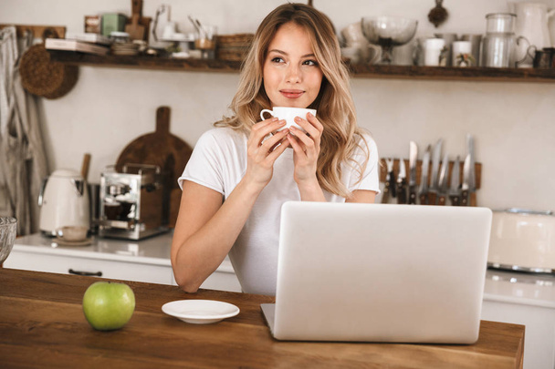 Image of caucasian blond woman drinking tea and using laptop whi - Фото, изображение
