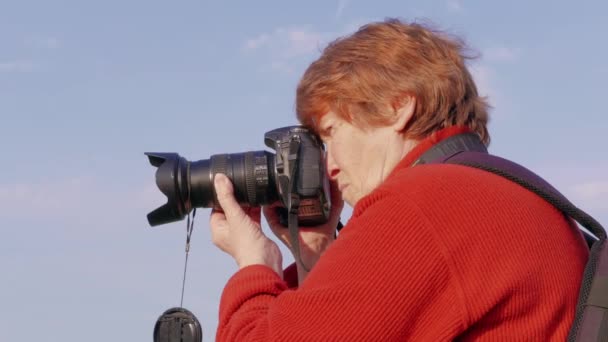 Senior nő fotózásra DSLR fényképezőgép, élességállítás. Nagymama hobbi kültéri. Idős hölgy lövöldözés, mutató kamera, húz hangsúlyt. Nyugdíjas fotós véve kép kívül, kék eget. Nagymama bevonva éles élességállítás, technológia segítségével - Felvétel, videó
