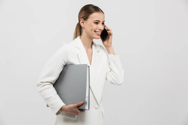 Beautiful amazing woman posing isolated over white wall background holding folder talking by mobile phone. - Photo, Image