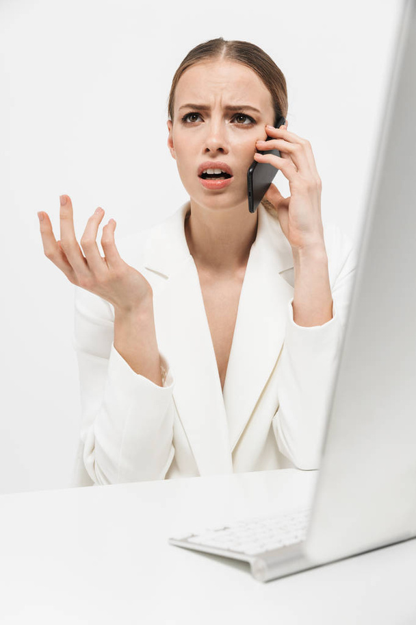 Displeased woman sitting isolated over white wall background using pc computer talking by mobile phone. - Valokuva, kuva