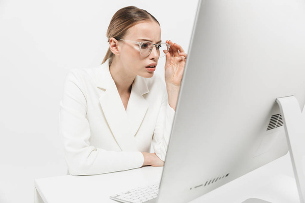 Όμορφη σοβαρή καταπληκτική γυναίκα κάθεται απομονωμένη πάνω από λευκό τοίχο φόντο χρησιμοποιώντας υπολογιστή PC. - Φωτογραφία, εικόνα