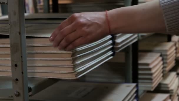 Uma mulher puxa para fora um livro grande ou jornal ajustado de uma prateleira da biblioteca. Mão de um bibliotecário de perto. De volta à escola. Formação e educação
. - Filmagem, Vídeo