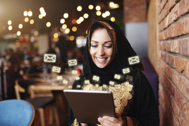 Gyönyörű muzulmán nő Toothy mosoly öltözött hagyományos viselet segítségével tabletta küldésére üzeneteket ülve kávézóban. - Fotó, kép