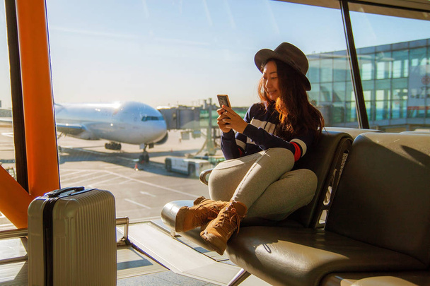 Young beautiful woman smiling, looking at mobile phone and chatting. Modern light airport before flight. - Photo, Image