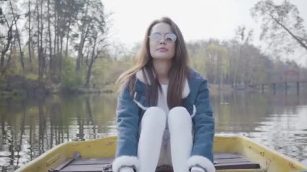 Portrait of a pretty young woman in sunglasses and a denim jacket floating on a boat on a lake or river. Beautiful brunette is actively relaxing on a day off or traveling enjoying nature. - Footage, Video