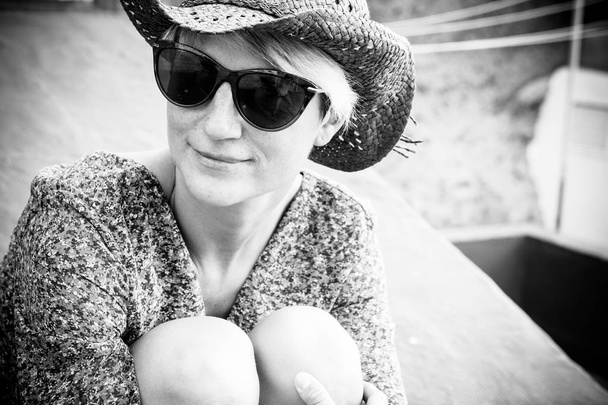 Beautiful black and white romantic portrait with blonde young caucasian woman sitting and hugging her nkees outdoor with sunglasses and little smile - Foto, Bild