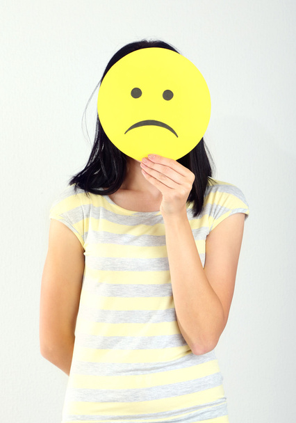 Young woman holding paper with sad smiley on gray background - Foto, Imagem