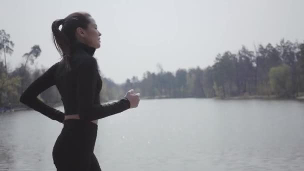 Attractive slim woman in sportswear running on the riverbank. Active lifestyle. sport. The lady keeping her body in shape. Beautiful landscape. Side view - Video